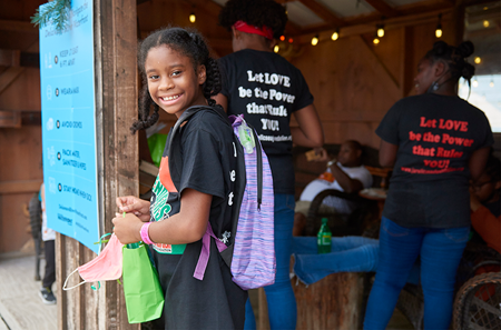 A young Black girl smiles at the camera while wearing a backpack and holding a small gift bag.